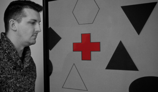 A photograph shows a person staring at a screen that displays one red cross toward the left side and numerous black and white shapes all over.