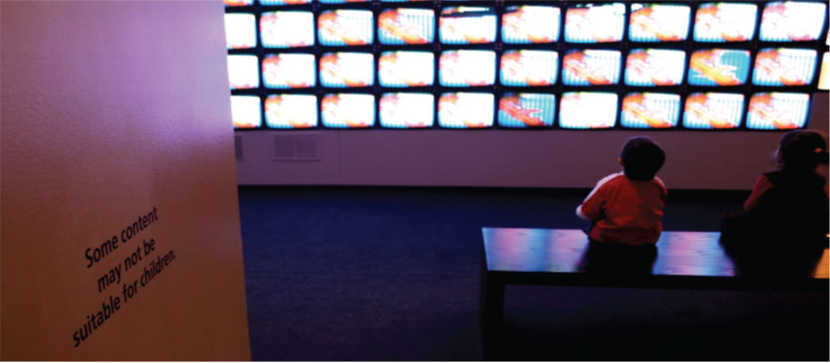 Children sit in front of a bank of television screens. A sign on the wall says, “Some content may not be suitable for children.”