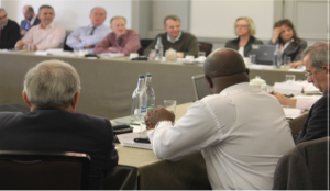 A photograph shows a group of people seated around tables in a meeting room.