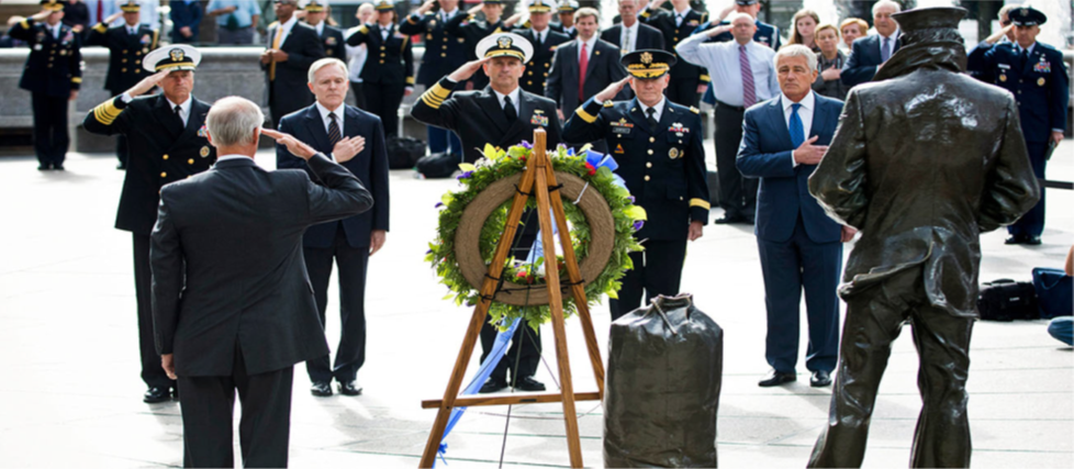 A photograph shows several key members of the United States military accompanied by a crowd as they stand facing toward a wreath. All hold their right arms in salute or placed across their chests.