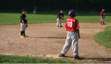 A photograph of children playing baseball is shown. Five children are in the picture, two on one team, and three on the other.