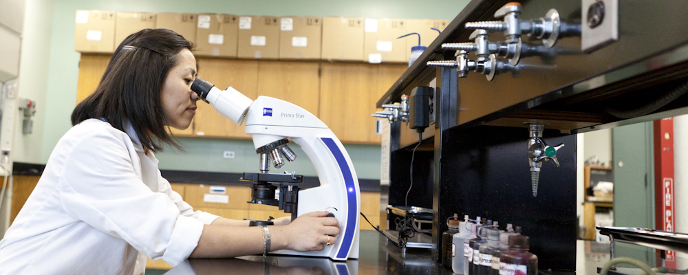 Student in lab coat looking at a slide under a microscope.