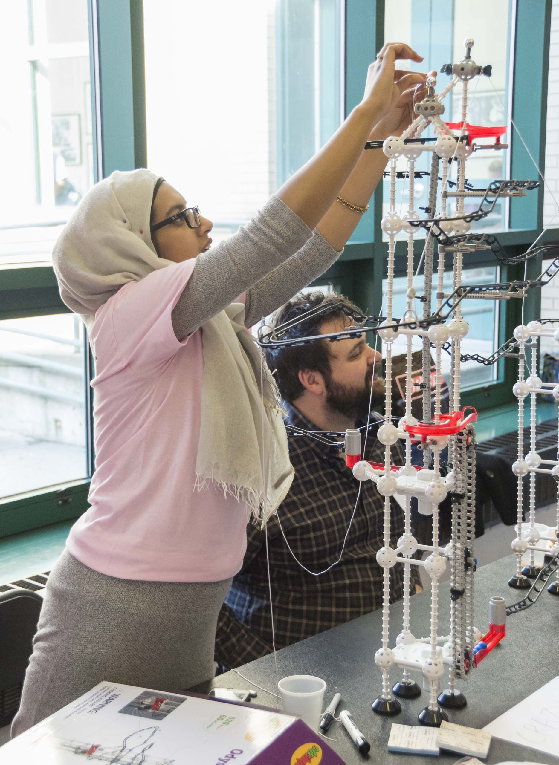 Student in headscarf building tower and bridge structures with toy connectors and adding weight to test integrity.