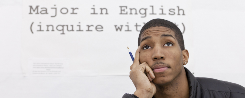 Student sits in front of a sign that says major in English inquire within.