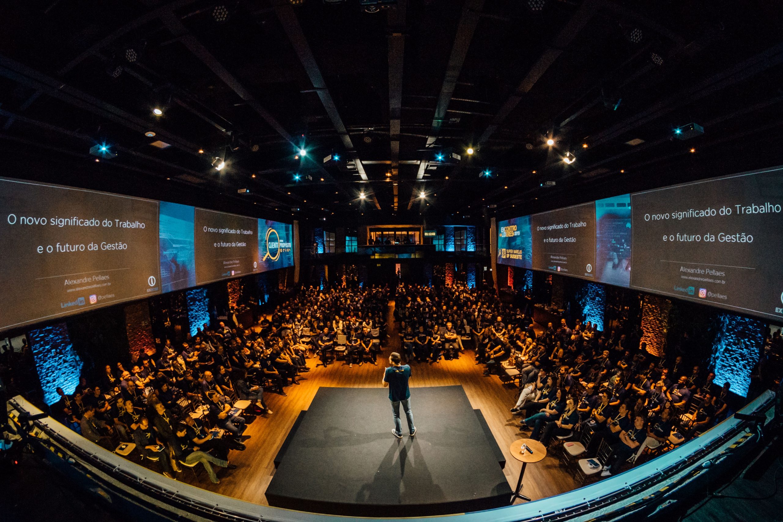 a speaker stands on the stage of an auditorium surrounded by large video screens and a full audience