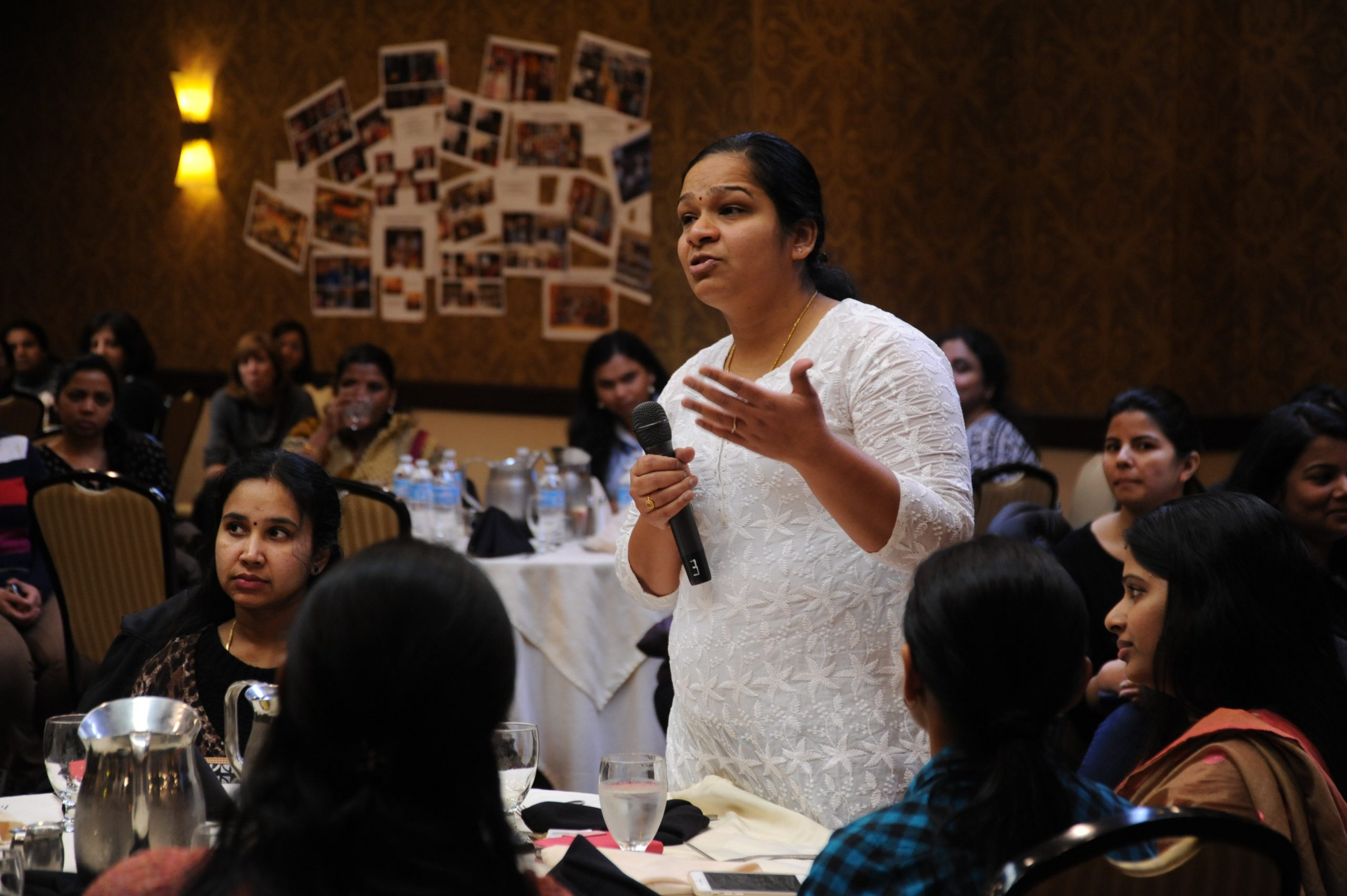 a South Asian woman standing and speaking into a microphone to an audience seated at dining tables