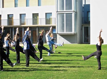 Photo shows a yoga instructor demonstrating a yoga pose while a group of students observes her and copies the pose.