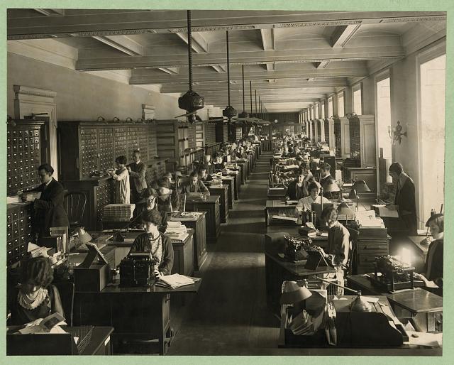 mostly female clerical workers at their desks with machines looking like typewriters, all in straight rows with a center aisle