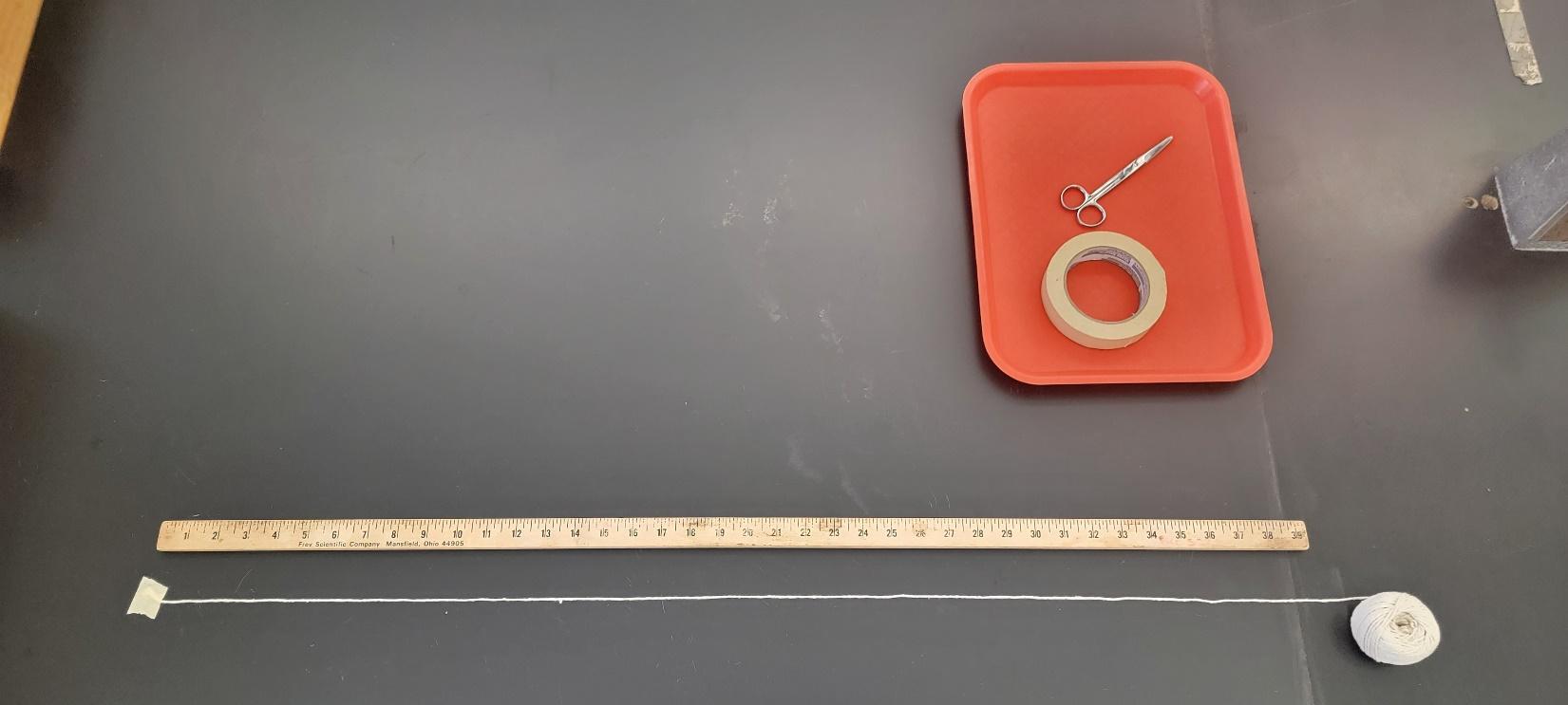 A wooden ruler lies parallel to a 4.6 meter length of white thread with Scotch tape attaching the loose end to the lab bench. Scissors and a roll of masking tape sit in a tray nearby.