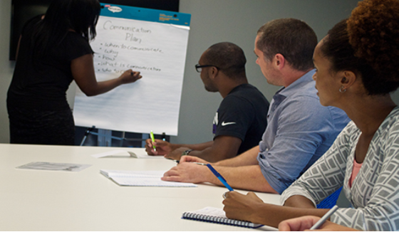 A photograph shows several people sitting at a table and writing on notepads, as a person in the front of the room writes on a large tablet.