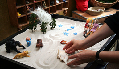 This photograph shows a person playing with objects in a small box filled with sand. The person is organizing these objects and small play figures in a form of treatment called sandplay.