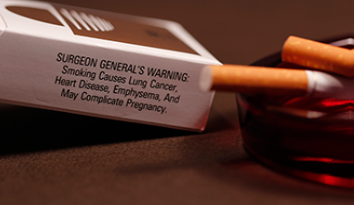 A photograph shows pack of cigarettes and cigarettes in an ashtray. The pack of cigarettes reads, “Surgeon general’s warning: smoking causes lung cancer, heart disease, emphysema, and may complicate pregnancy.”