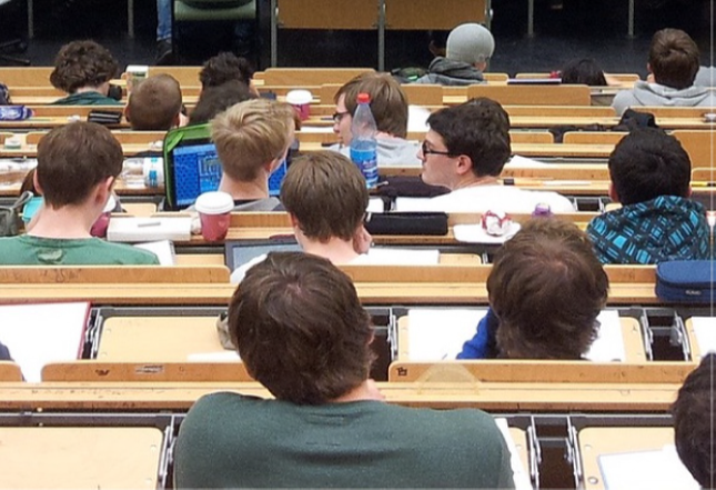 2.15 Many rows of students are in a classroom. One student has an open laptop on his desk.