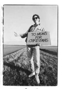Black and white photo of a white man holding a hand-painted sign with "To Mom's for Christmas" in all caps, right thumb up, along the side of a road in a grassy plain